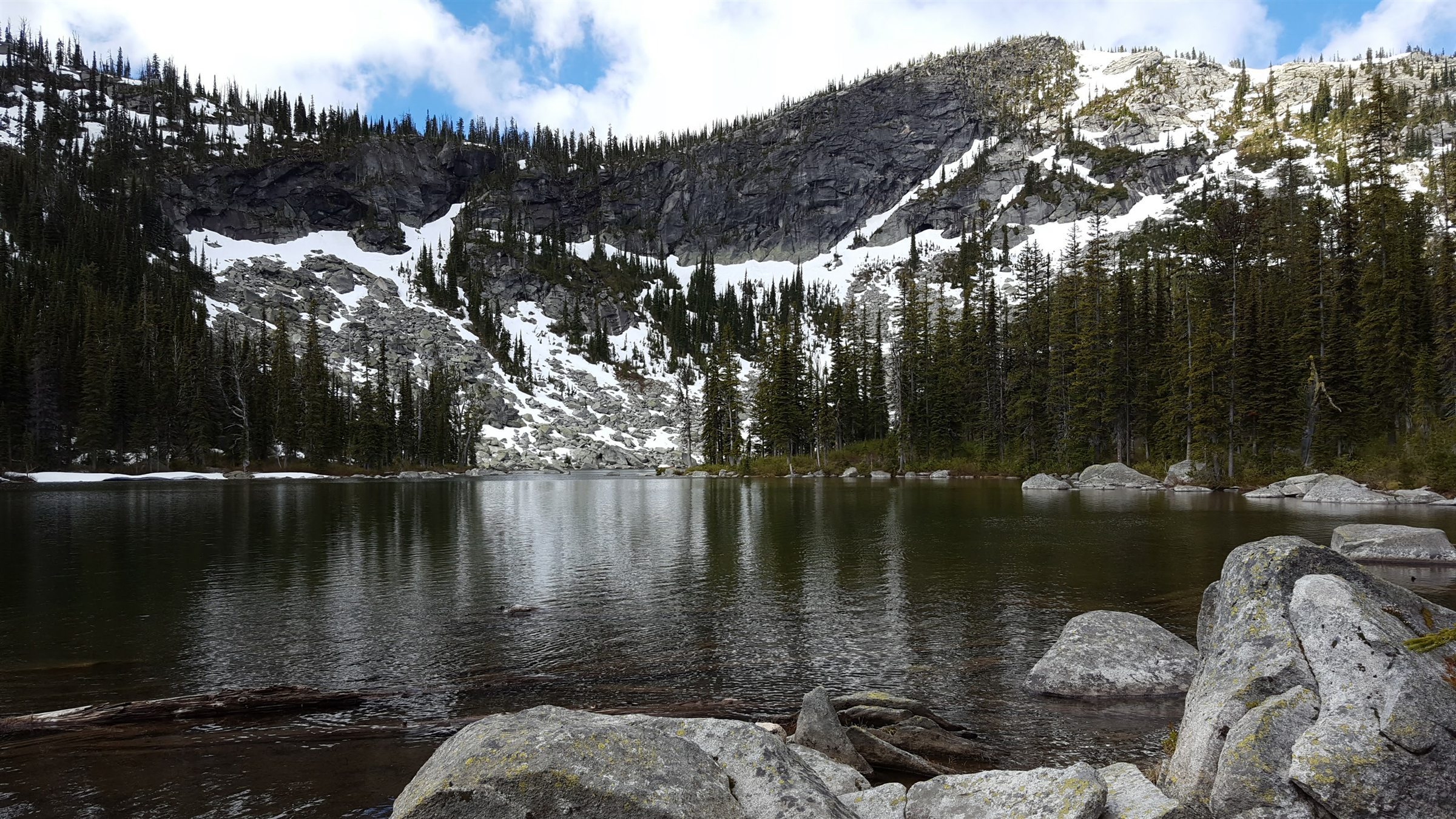A still alpine lake below snow-covered rock walls and evergreen trees