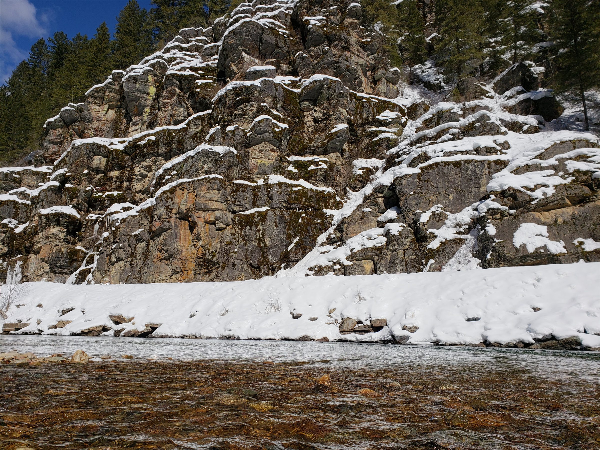 A snow-covered rock wall rising above a clear winter river