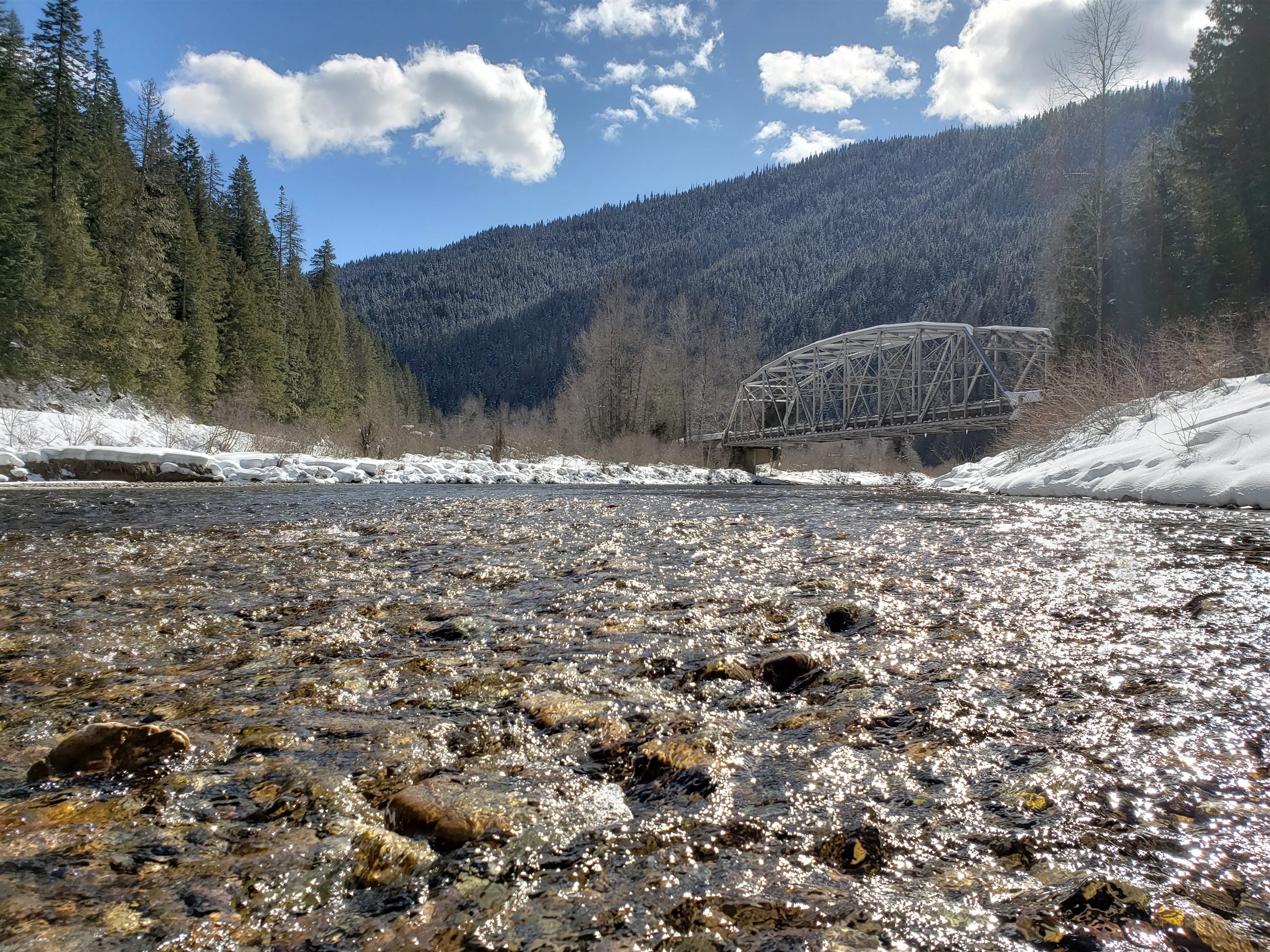A bright winter river flowing toward a steel bridge between snowy mountains